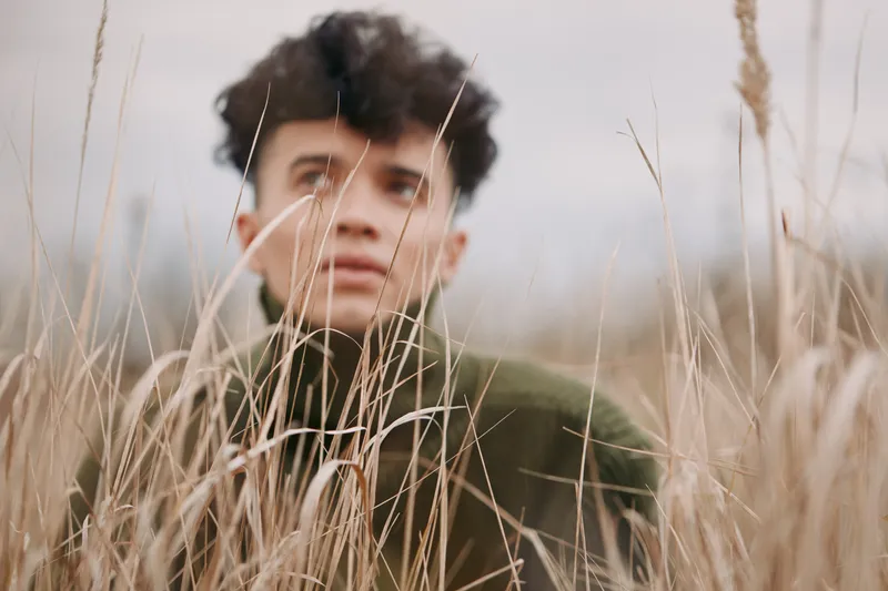 Candid portrait of a young person peering through tall dried grass, conveying authenticity, introspection and quiet resilience in a muted outdoor scene with genuine natural emotion.