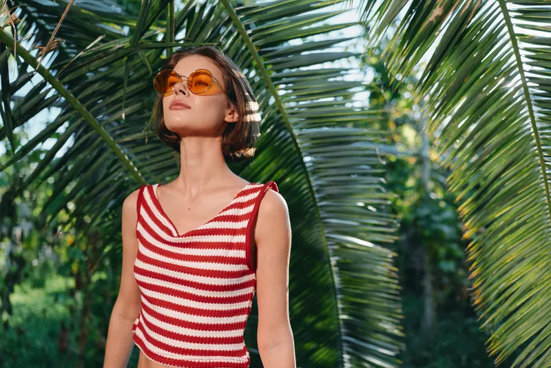 A stylish woman stands in a tropical garden wearing a red striped sleeveless top and orange sunglasses, basking in sunlight with a confident, carefree vibe and summer allure.