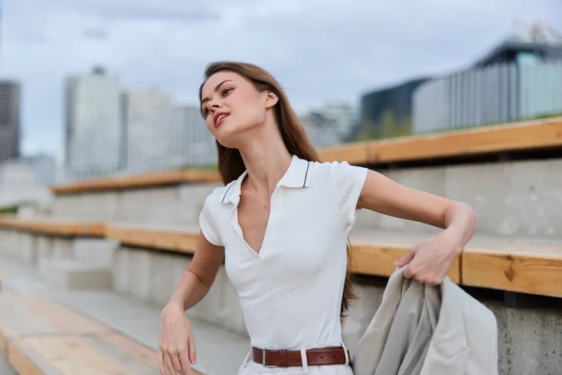 Young woman in white shirt outdoors in urban environment with confident expression and beige jacket in hand, casual fashion style, summer season