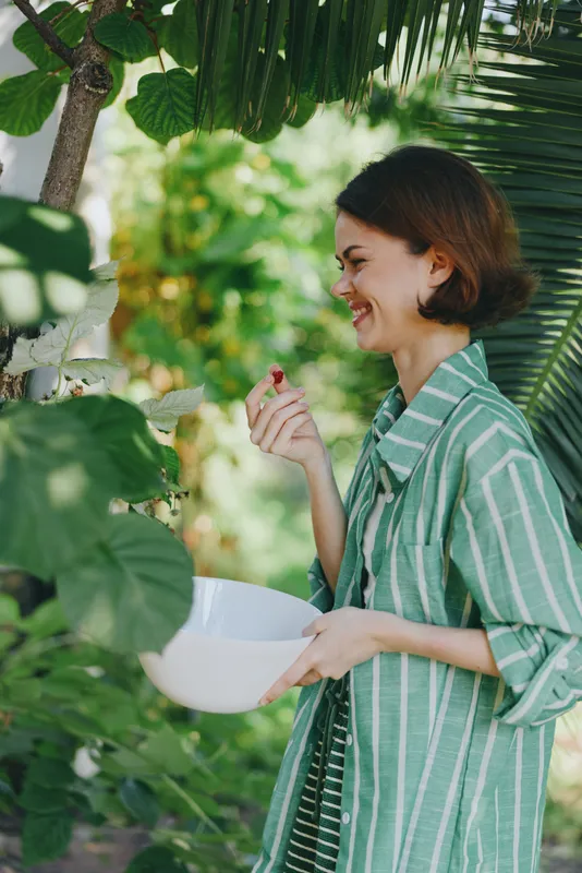 Woman smiling outdoors while holding a bowl, standing among lush garden greenery, wearing a striped green shirt, capturing a moment of simple joy, calm nature, and fresh air.