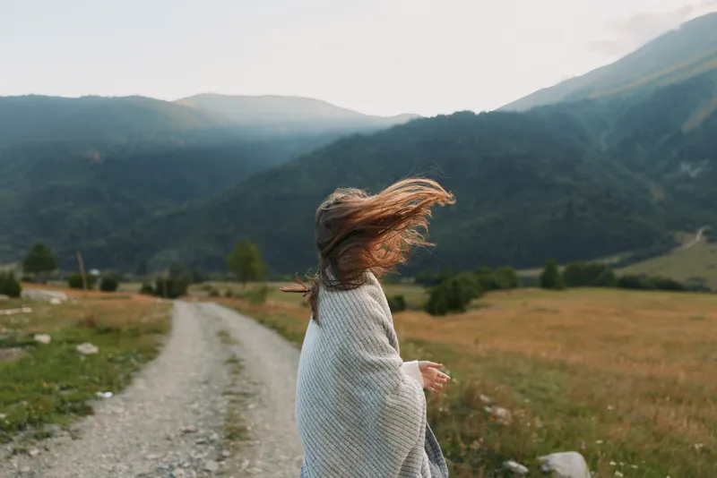 A woman wrapped in a blanket stands on a gravel path in a mountain valley during sunset, her hair blowing in the wind. The scene reflects peaceful lifestyle and nature connection.