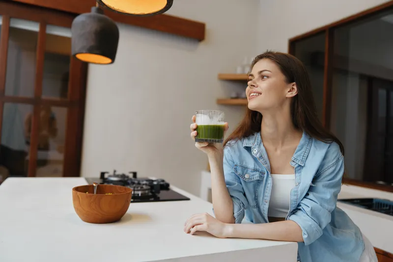 Веселая молодая женщина держит стакан зеленого смузи в современной кухне, одетая в повседневную джинсовую куртку, демонстрируя яркий и здоровый образ жизни