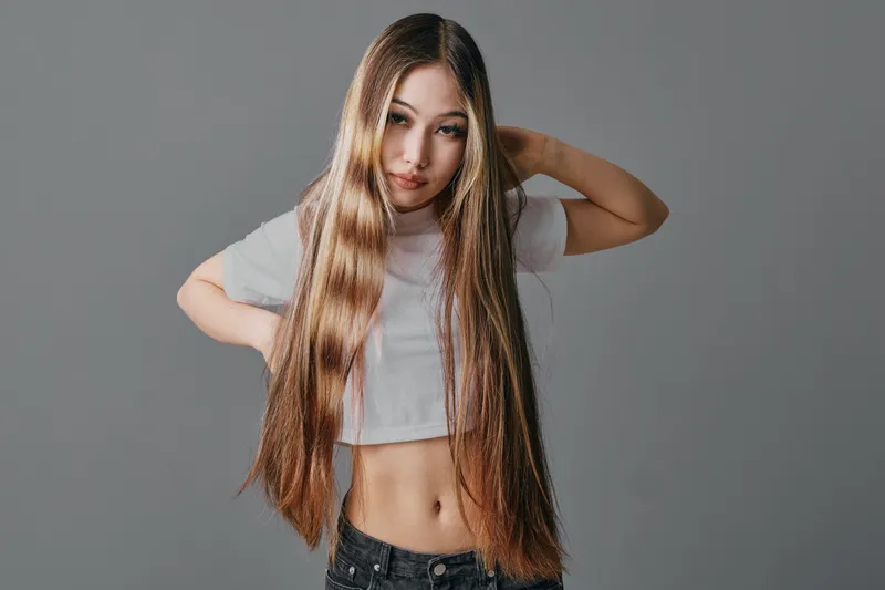 Teenage girl with long straight hair wearing a white crop top and black jeans posing against a gray studio background looking at camera with hand behind head.