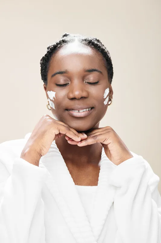 Smiling Black woman applying skincare minimalist routine in soft light, showcasing natural beauty and self care with facial cream