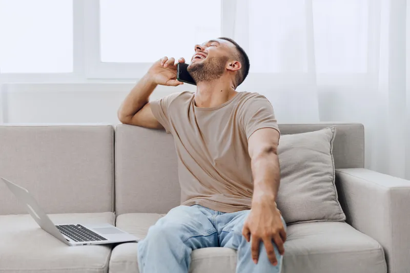 Joyful man talking on the phone while sitting on a cozy couch, embodying the comfort of remote work and modern living in a bright, airy space.