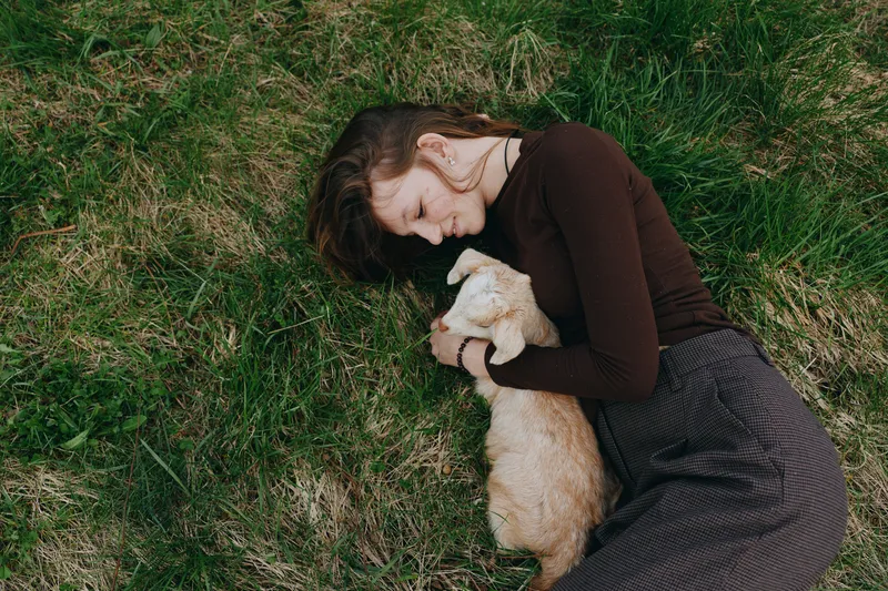 Young woman rests on grass outdoors, embraces a small dog with tender affection, a calm cozy moment captured in natural light, depicting a simple lifestyle of companionship