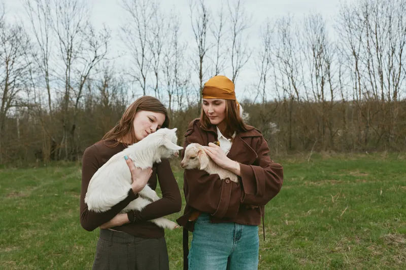 Two women outdoors in a grassy field, smiling while holding playful goats, warm coats and a soft beanie, candid lifestyle moment, natural light, calm countryside ambiance, gentle connection with