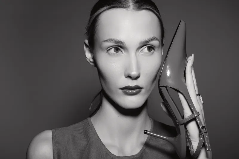 A slim white woman with pale skin holds a shiny high heel shoe beside her face in a creative black and white studio portrait, fashion and style concept.