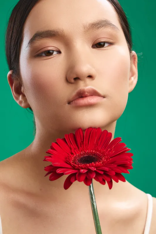 Asian woman with a calm expression holds a red flower near her chin against a vibrant green background, creating a minimalist, elegant portrait.