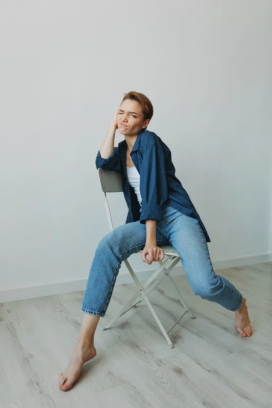 Молодая женщина сидит дома в кресле, улыбается, показывая зубы, с короткой стрижкой, в джинсах и джинсовой рубашке на белом фоне
