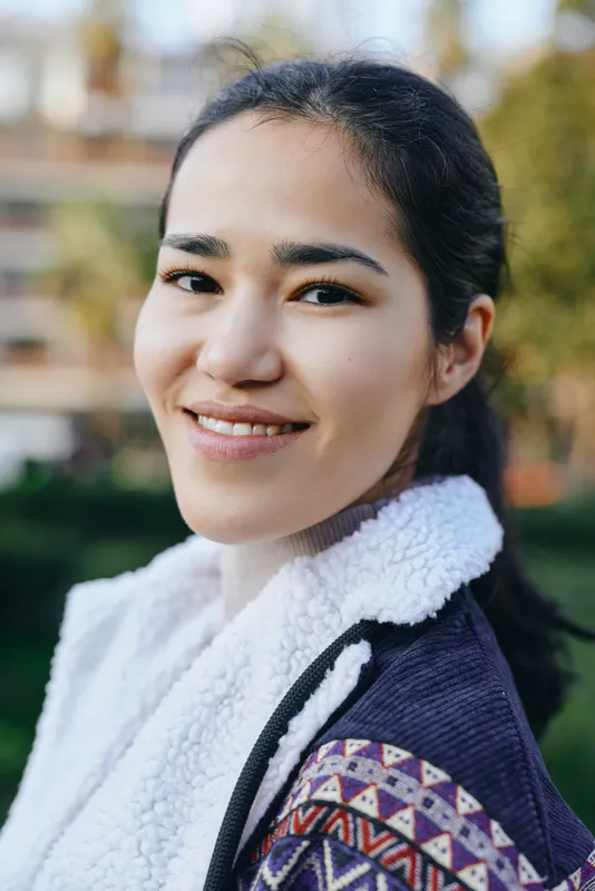 Woman with a warm smile in an outdoor setting, wearing a patterned jacket and fluffy scarf, close to camera with soft background and natural light, relaxed lifestyle fashion image for travel, daily