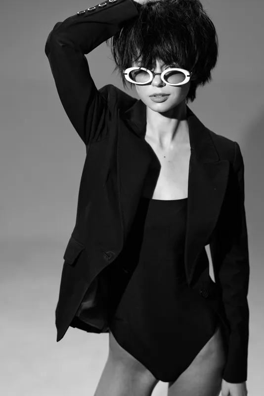 Stylish woman wearing sunglasses and a black blazer posing confidently in black and white lighting.