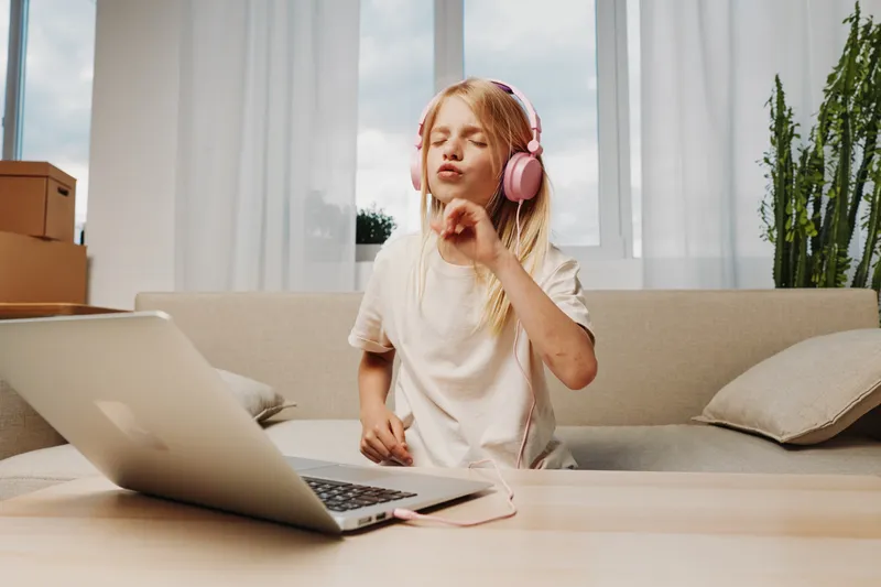 Blonde girl with pink headphones enjoys music with closed eyes, sitting on beige couch near laptop in bright living room. Concept of relaxation, childhood, technology, and leisure time.