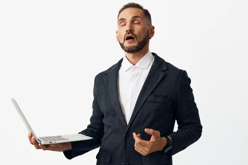 A professional businessman stands against a white background, holding a laptop in one hand and gesturing with the other, expressing astonishment and confident emotion during a work moment.