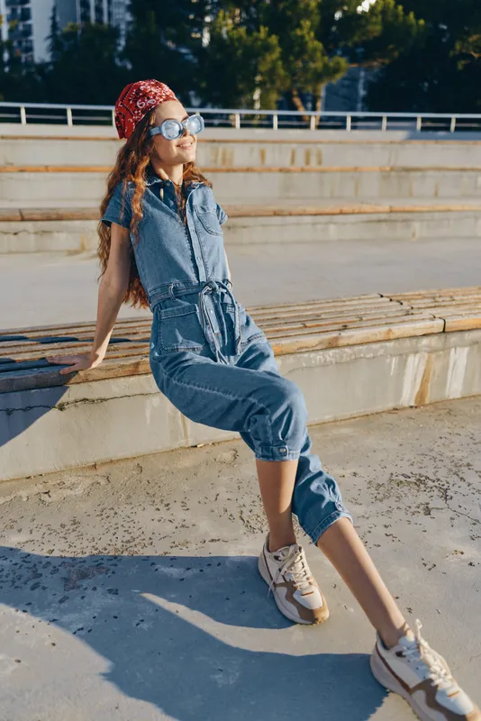 young woman wearing sunglasses and denim jumpsuit relaxing outdoors on sunny day in urban setting with confident and carefree expression