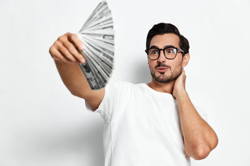Young man holding fan of money, looking thoughtfully at cash, wearing casual white t-shirt and glasses, isolated on plain solid background. Finance concept.