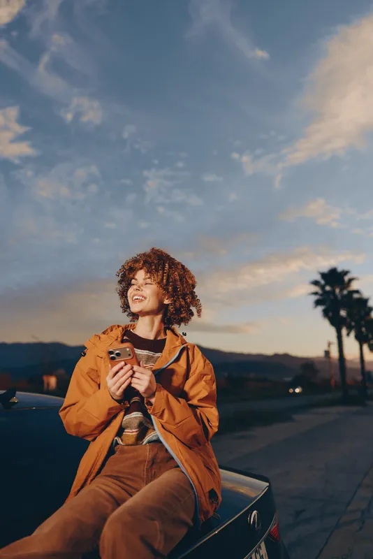 Woman lifestyle outdoors at sunset, curly haired woman in orange jacket, relaxed on car hood, holding coffee cup, warm evening light, calm smile, palm trees background, soft clouds