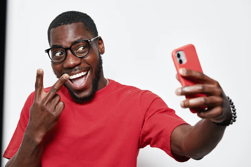 Black man with dark skin wearing glasses and red shirt taking selfie with smartphone showing peace sign and smiling in studio with white background.