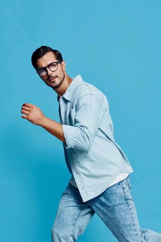 Full body smiling man in casual light blue shirt and glasses looking confidently at the camera against a solid blue background. Fashion lifestyle concept.