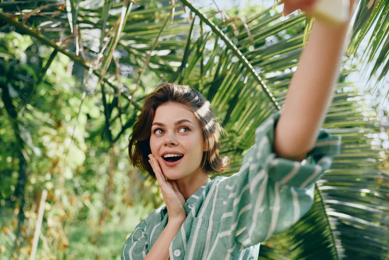A cheerful woman outdoors raises her arm and smiles, enjoying a sunny day among lush greenery.