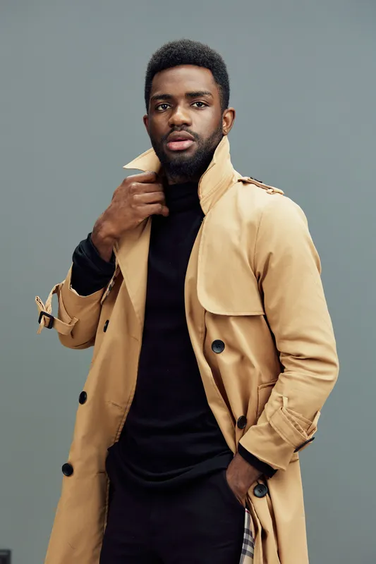 Stylish black man in a beige trench coat and turtleneck sweater, showcasing confidence and modern fashion against a muted gray background