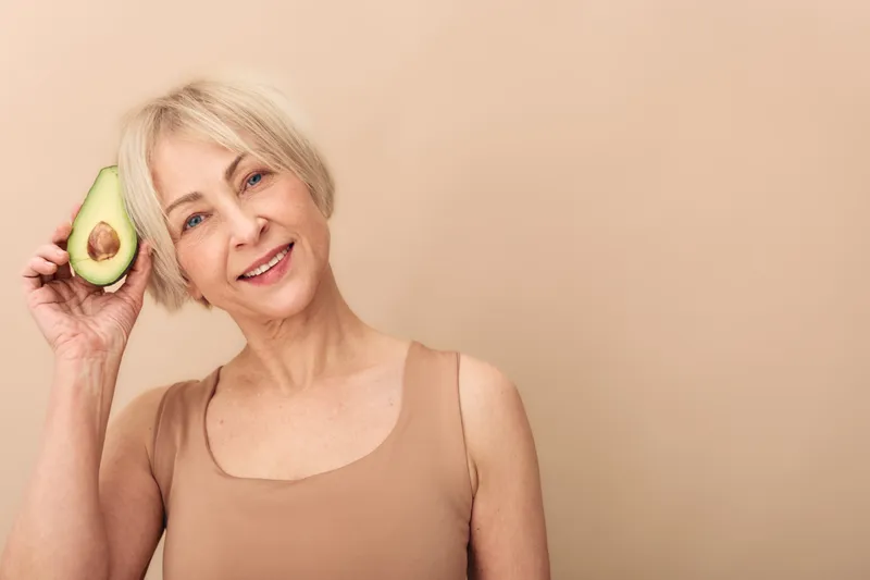 Smiling mature woman with short blonde hair holds half avocado near head, wearing beige tank top. Concept of healthy lifestyle, nutrition, natural beauty, and wellness in studio with neutral