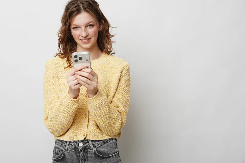 Smiling young woman in yellow sweater holds smartphone with both hands, looking at camera. Casual style, communication, technology, and positive mood on light background.