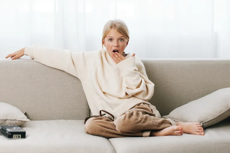 A young girl sits barefoot on a beige sofa in a cozy living room, surprised with a hand to her mouth while wearing a soft cream sweater.