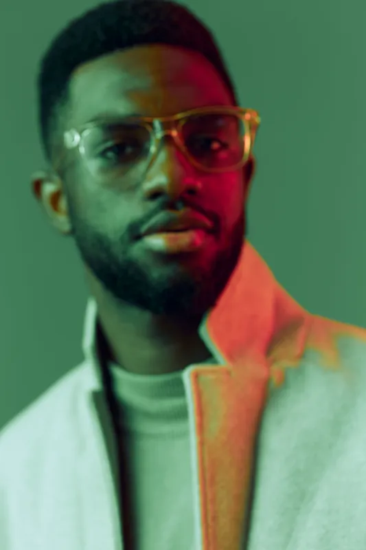Stylish young man with glasses in a trendy outfit, showcasing confidence against a soft green background with a hint of pink light