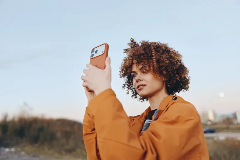 Woman selfie outdoors lifestyle portrait curly hair orange jacket, holding phone up for photo, smiling and looking at screen, daytime sky background, casual city walk, modern technology