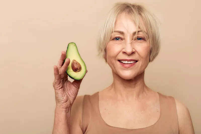 Smiling mature woman holds half avocado near face on beige background. Concept of healthy lifestyle, natural food, skincare, nutrition, and wellness for older adults.