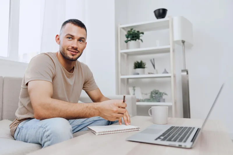 Smiling man sitting on a couch, engaged in creative work with a notebook and laptop in a modern, bright home office environment. Fresh and inviting atmosphere.