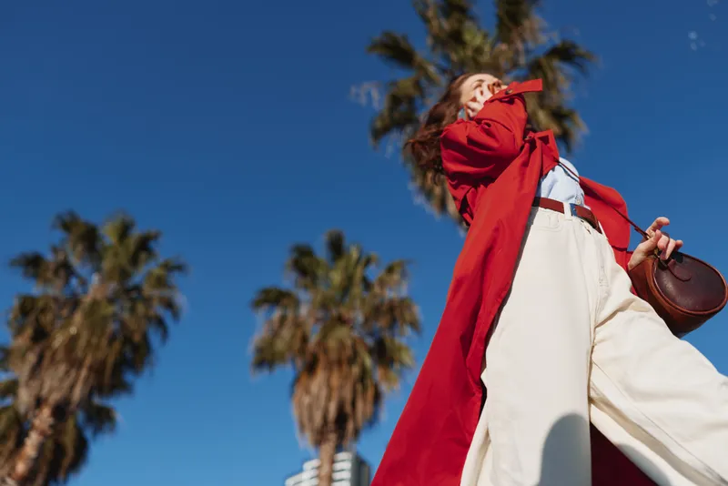 Young woman in a red coat and white pants strides under palm trees against a bright blue sky, exuding bold confidence and playful movement as her hair flows freely.