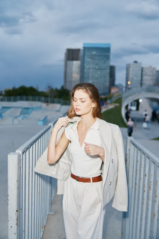 Young stylish woman in white suit walking with city skyscrapers background during cloudy day, confident urban business fashion concept