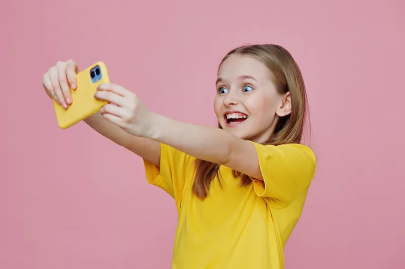 A cheerful young girl with long hair taking a selfie with a smartphone against a pink background, wearing a bright yellow t shirt, expressing happiness and joy