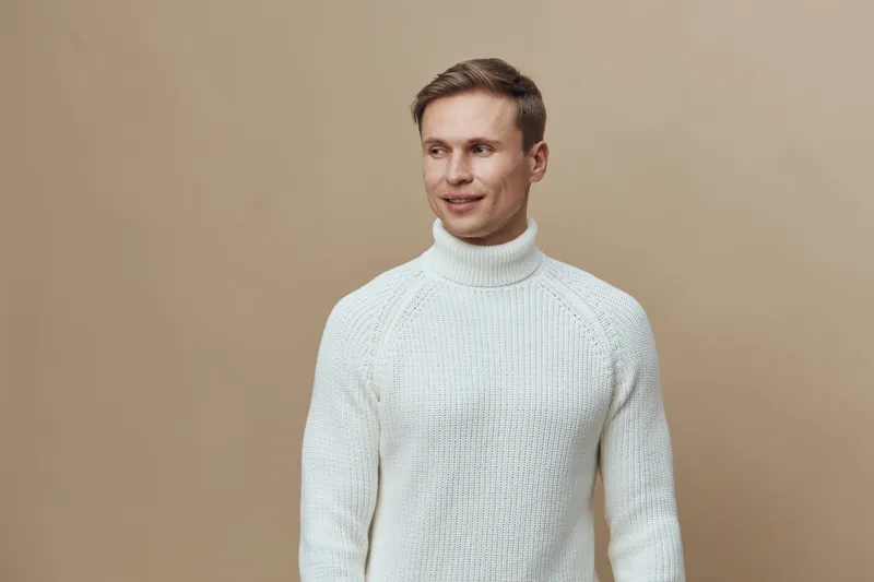 Smiling man wearing white turtleneck sweater looking to the side on plain beige background studio portrait. People lifestyle concept