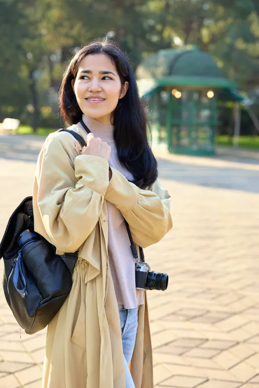 Woman wearing a beige coat and pink sweater with a backpack and camera, standing in a sunlit park, exploring outdoors, casual style, candid lifestyle, joyful expression, travel vibe, natural light,