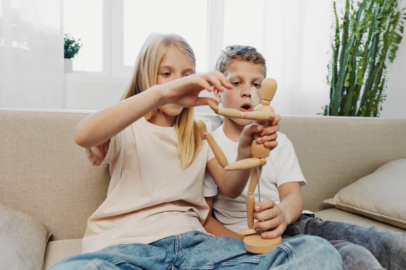 Two children sitting on sofa play with wooden mannequin, exploring creativity and imagination in a bright living room with natural light and casual clothes. Concept for childhood play and learning.