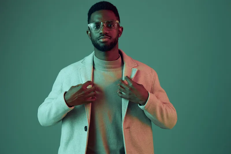 Confident African man in stylish coat and sweater, posing against a muted green background, embodying modern fashion and individuality