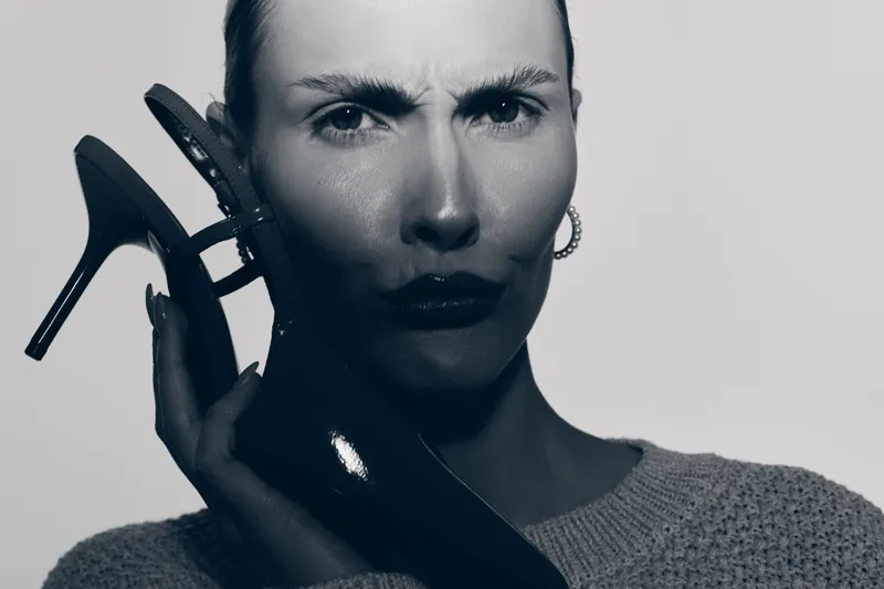 A serious white woman with pale skin holds a black high heel shoe near her face in soft light. Concept of fashion, style, footwear, and emotional expression.