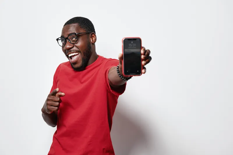 African American man with dark skin wearing glasses and red shirt smiling and showing smartphone screen while pointing finger on white background.