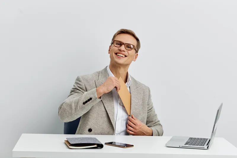 cheerful smiling man wearing grey jacket holding envelope sitting at white desk with laptop and smartphone digital nomad remote work concept