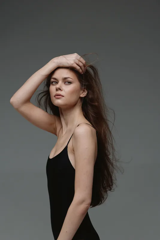 Young woman with long hair posing in black outfit with confident expression in simple studio setting.