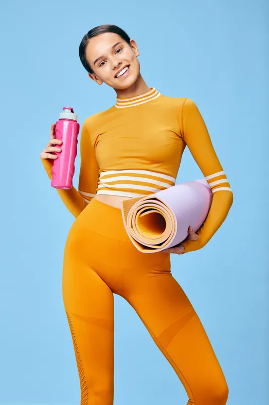 Young woman fitness sport scene: woman in orange activewear holds a rolled pastel yoga mat and pink water bottle against a blue studio background, smiling confidently for wellness lifestyle.