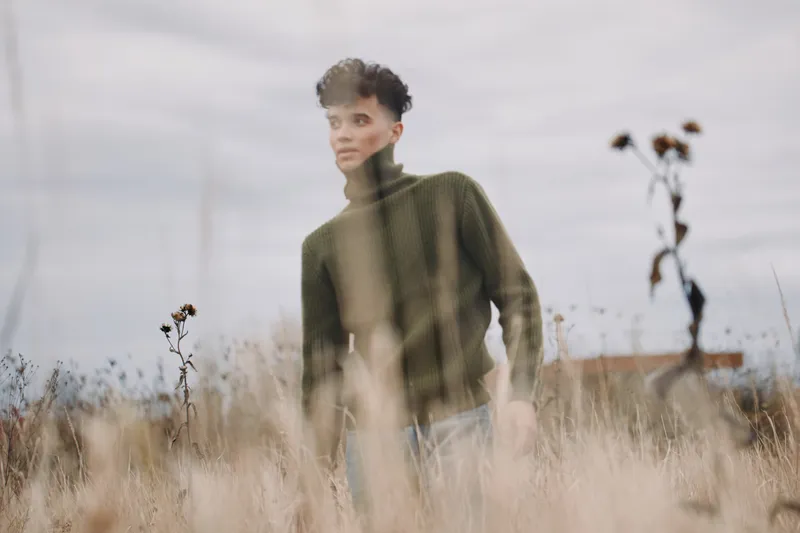 A young man stands in a muted meadow wearing a green turtleneck, photographed with authentic, natural expression and visual credibility, creating a candid, contemplative portrait in soft light.