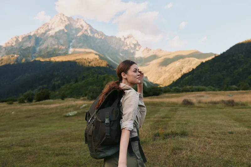 A woman hiker with a large backpack stands in an open meadow, looking toward distant mountains. Bright natural light captures exploration, resilience, and hopeful wanderlust.