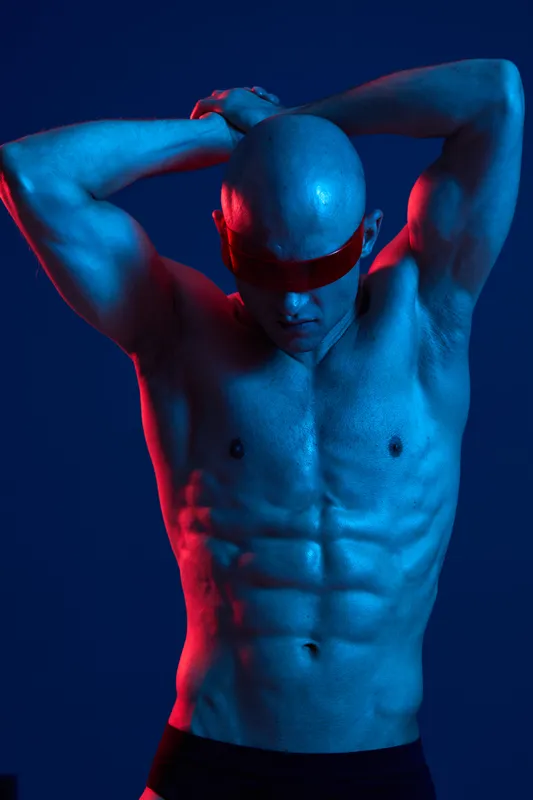 Strong man posing in dramatic lighting, showcasing his muscular physique and intense expression. Colorful blue and red hues enhance the visual impact of his physique.
