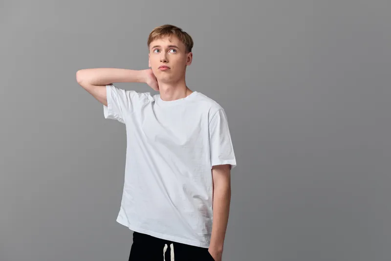 Casual man poses thoughtfully with hand at the neck against a plain studio backdrop, wearing a white t shirt and relaxed stance for a contemplative everyday look.