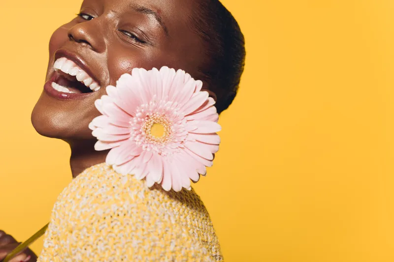 Happy smiling black woman with dark skin holds pink gerbera flower near face against yellow background. Concept of joy, beauty, natural skincare, and positive mood in studio lighting.