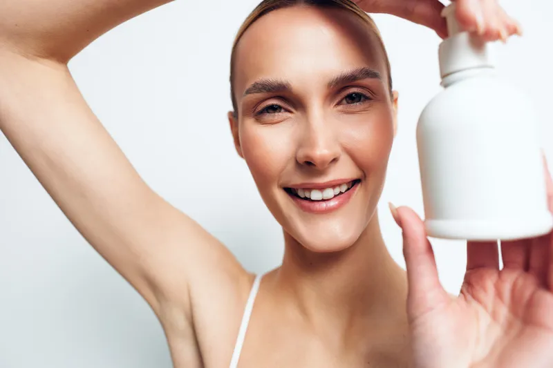 Woman smiles in a bright studio holding a skincare bottle and cosmetic serum, showing healthy self care. Clean background, soft light, arms raised, beauty routine.